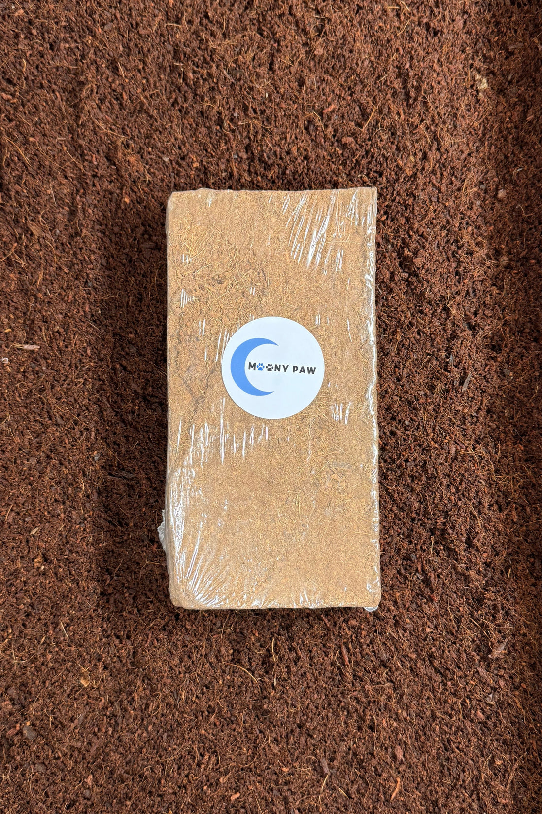 A compressed coconut fiber brick wrapped in plastic with a Moony Paw logo, placed on loose coconut soil.