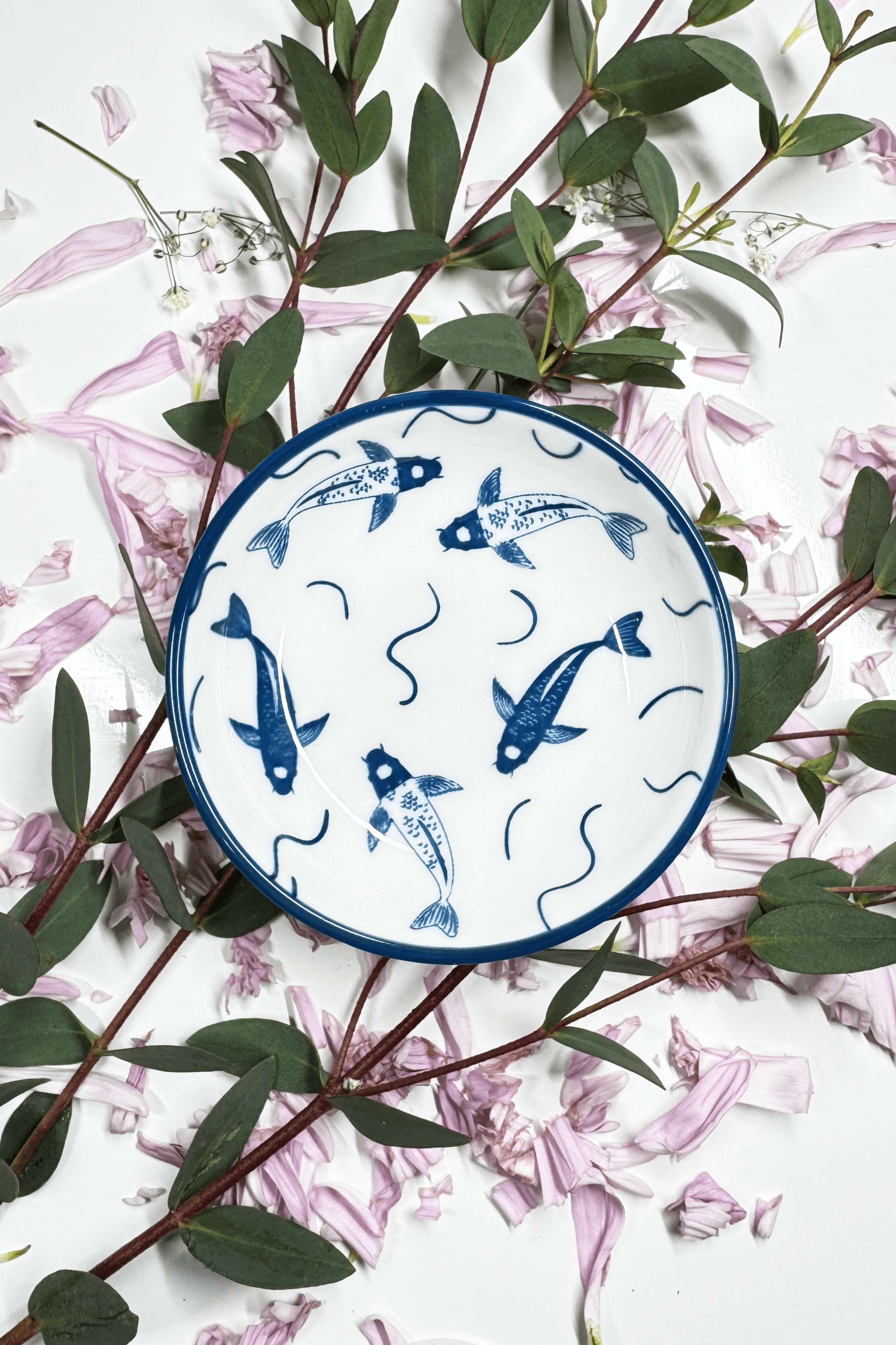 A Sapphire Splash Plate for small animals, featuring a blue koi fish design, placed on petals and greenery.