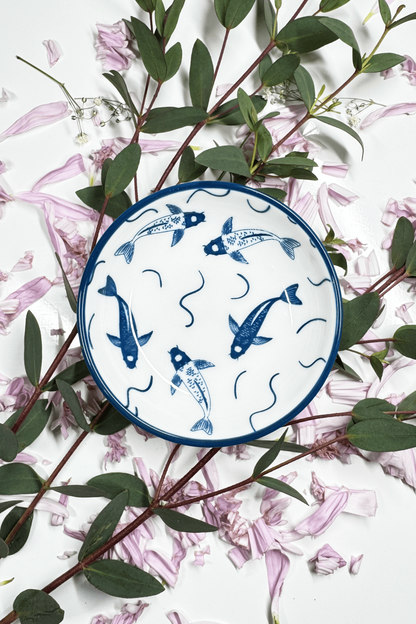 A Sapphire Splash Plate for small animals, featuring a blue koi fish design, placed on petals and greenery.